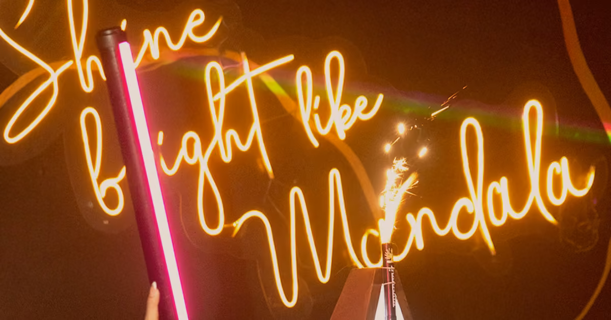 A neon sign reads Shine bright like Mandala. In front, a hand holds a pink light stick near a small sparkler on a pyramid.