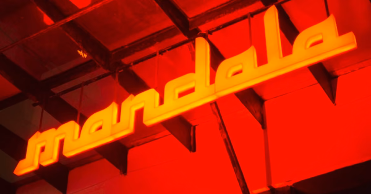 A glowing yellow neon "mandala" sign hangs on a red ceiling, casting a vibrant ambiance in the Mandala Night Club.