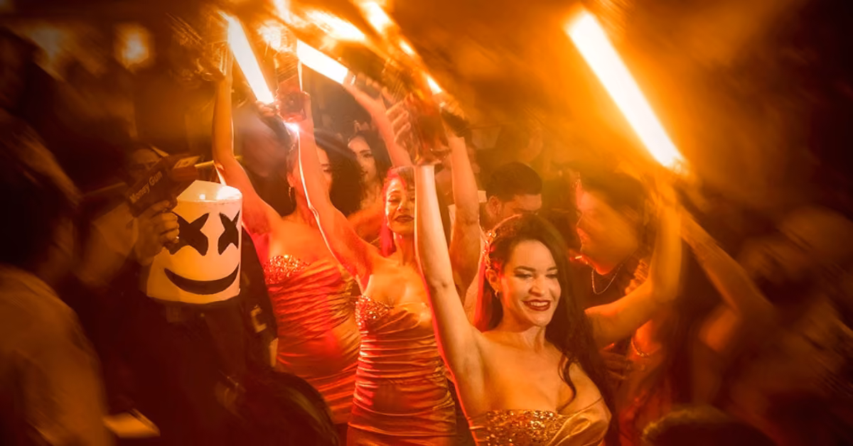 Women in gold dresses hold sparklers above their heads at a crowded party, with orange lights and energetic, blurred motion.