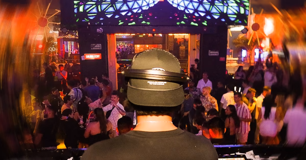 A DJ with headphones plays at Mandala Night Club as colorful lights and screens shine over a lively, dancing crowd.