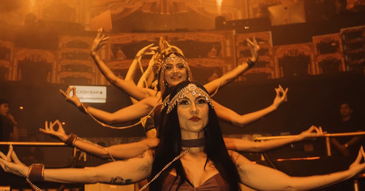 Women in ornate costumes and headpieces perform a synchronized dance in a line, creating an illusion of many arms on stage.
