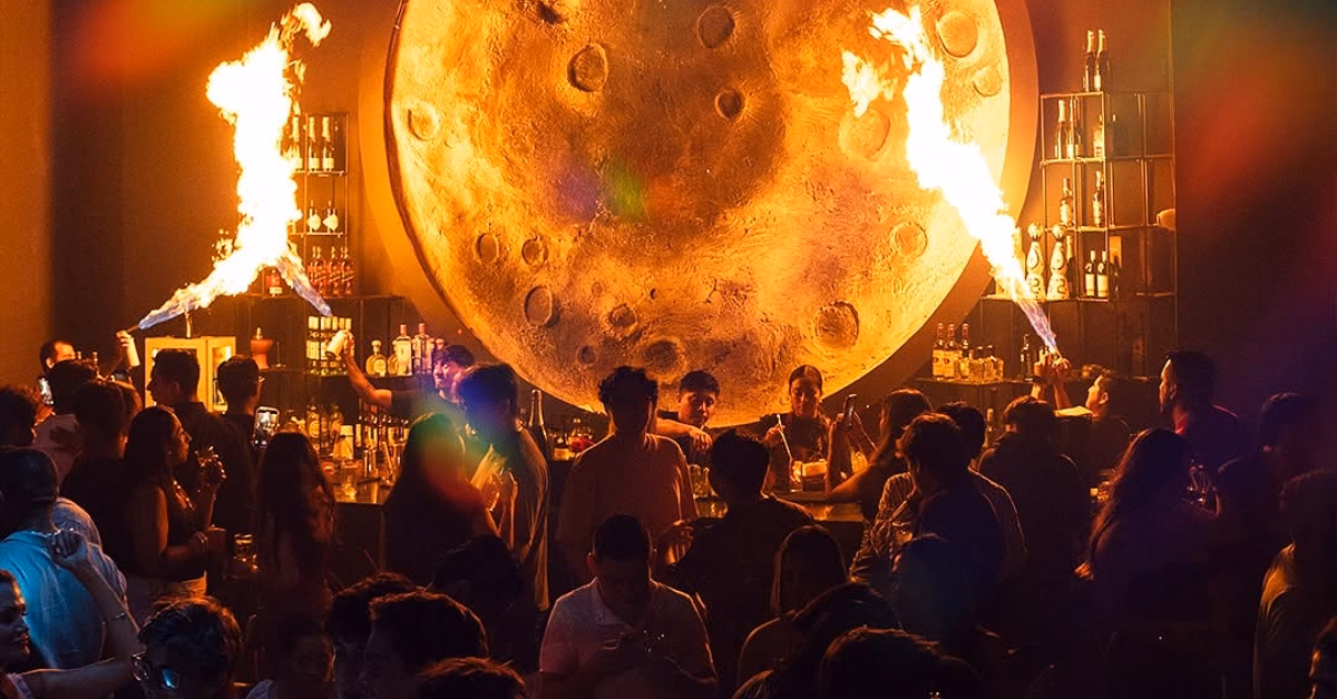 Lively bar with a moon backdrop, bottle-lined shelves, and fire displays. People socialize in vibrant, dim lighting at Mun Night Club.