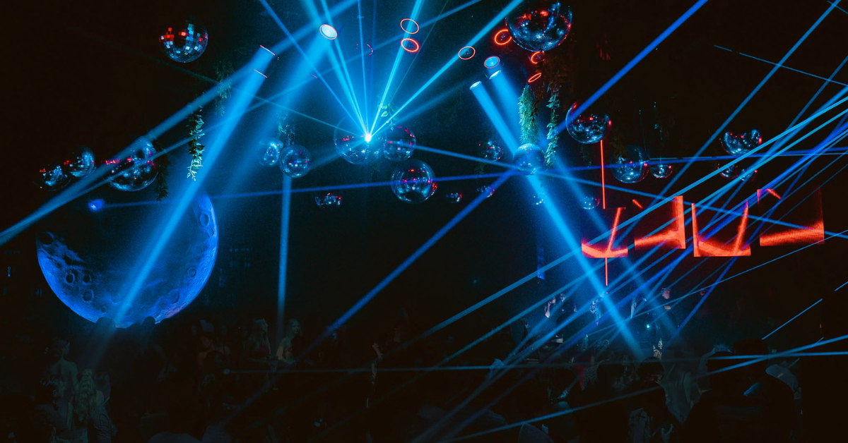 A dimly lit nightclub with blue laser lights, disco balls, and a dancing crowd. Red neon letters and a moon hang in back.