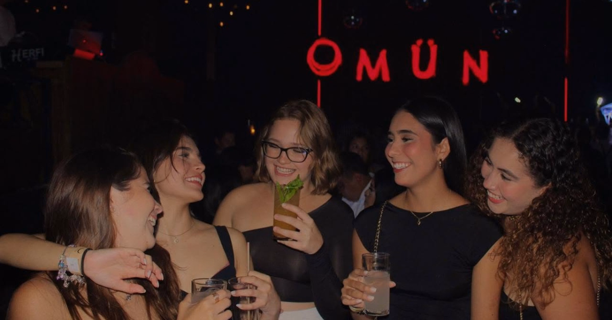Five women in black outfits smile and hold drinks at Mun Night Club, with a red neon OMUN sign glowing in the dim background.