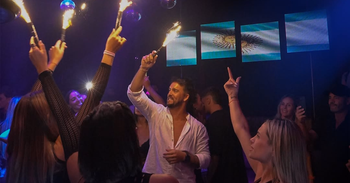 People celebrate at a nightclub with sparklers, smiling and cheering as screens in the background show the Argentine flag.