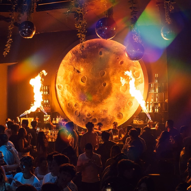 A lively nightclub with a glowing moon backdrop, fire effects, disco balls, and colorful lights shining over dancing partygoers.