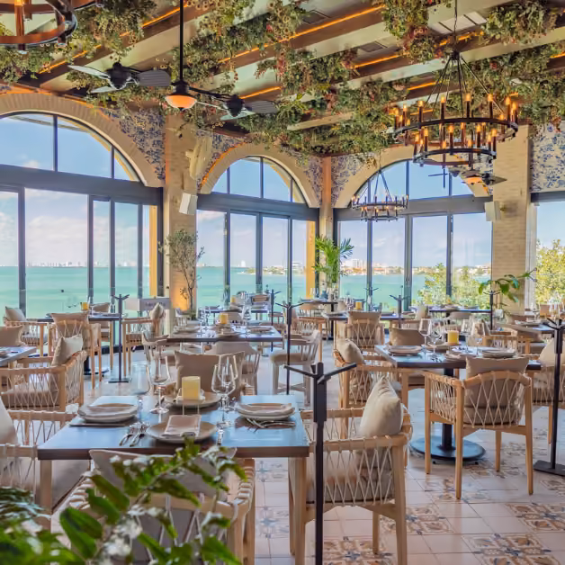 Elegant restaurant with arched windows facing the ocean, hanging plants, chandeliers, and neatly set tables with glassware.