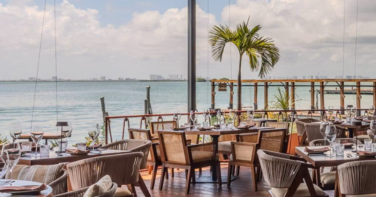 Seaside outdoor dining with wooden tables, chairs, and palm tree overlooking calm water and a distant city skyline.