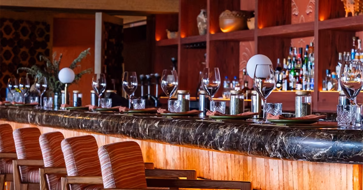 Porfirio’s bar with a marble counter, wooden chairs, set tables with wine glasses, and shelves of bottles in warm light.