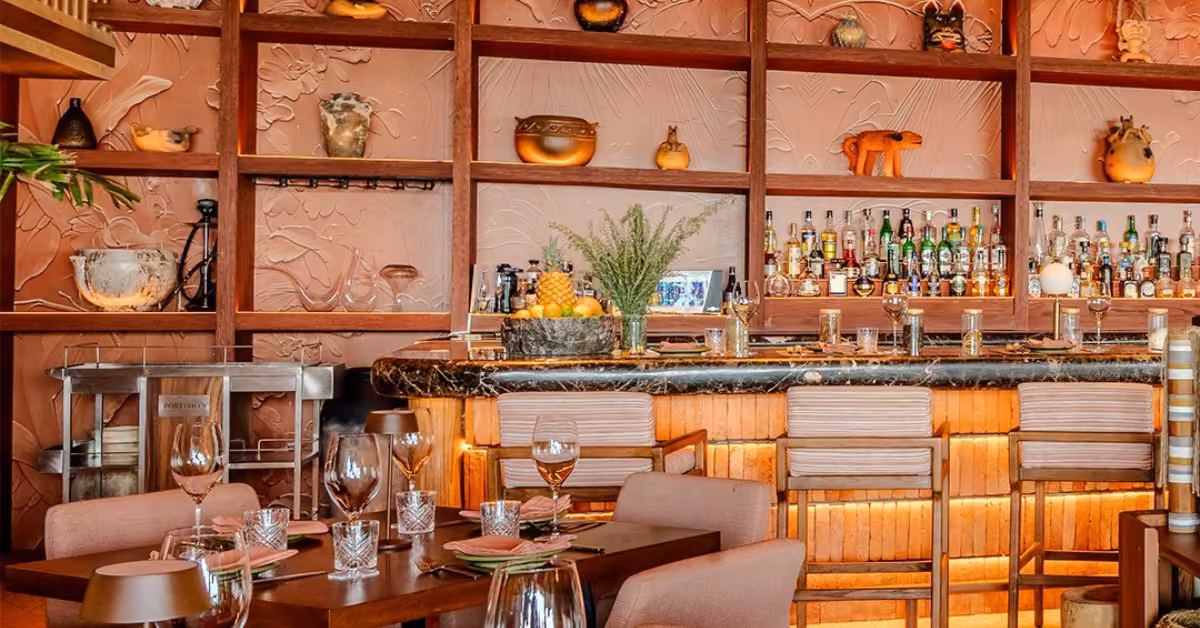 Porfirio’s bar with wooden shelves, pottery, bottles, marble counter, high stools, and warmly lit dining tables in front.