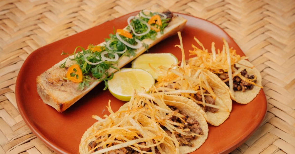 Three beef tacos with crispy potato strips, lime halves, and roasted bone marrow with herbs on a woven mat.