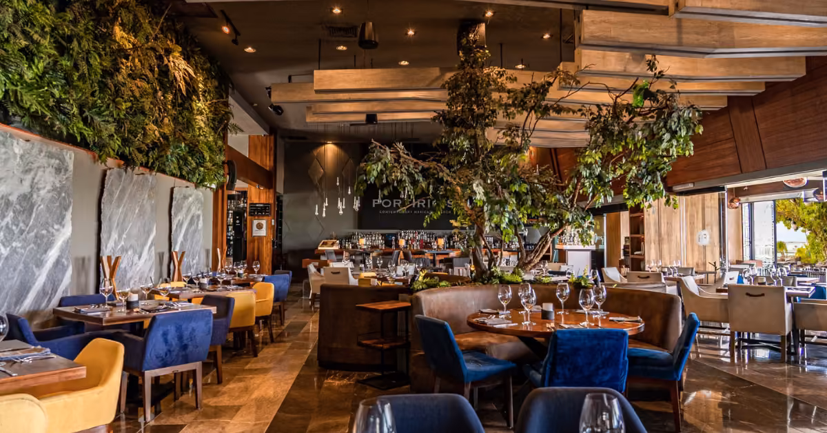 Porfirio’s Restaurant interior with elegant tables, blue-beige chairs, lush greenery, wood accents, and a central tree.