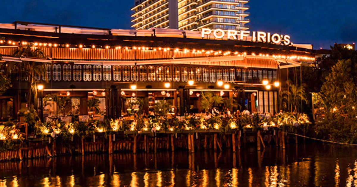 Porfirio’s Restaurant glows at night by the water, string lights shining as people dine outdoors with a tall building behind.