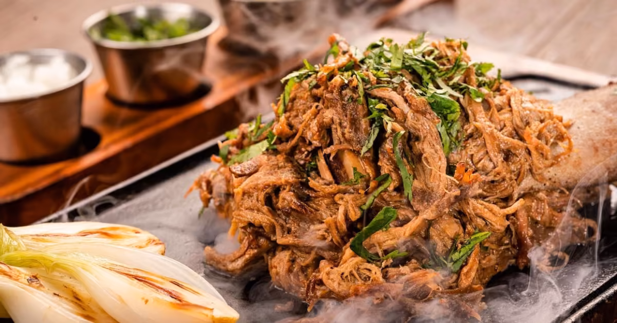 Shredded, seasoned meat topped with herbs sits beside grilled onions, with sauces and greens in the background.