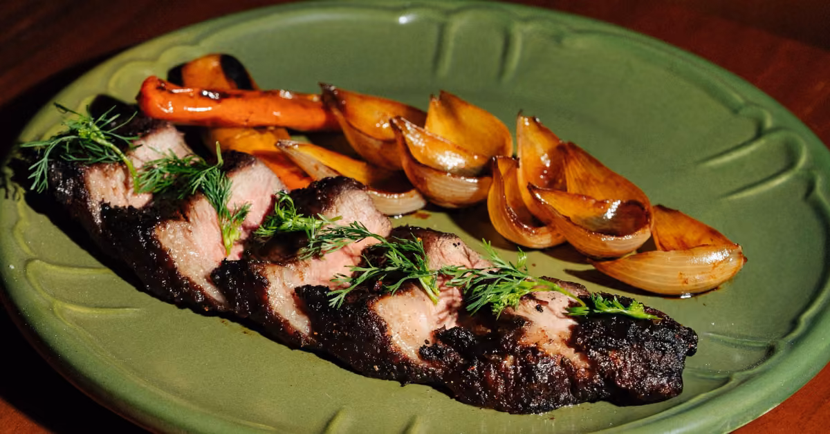 Sliced grilled steak with fresh herbs, served alongside roasted carrots and caramelized onions on a green plate.