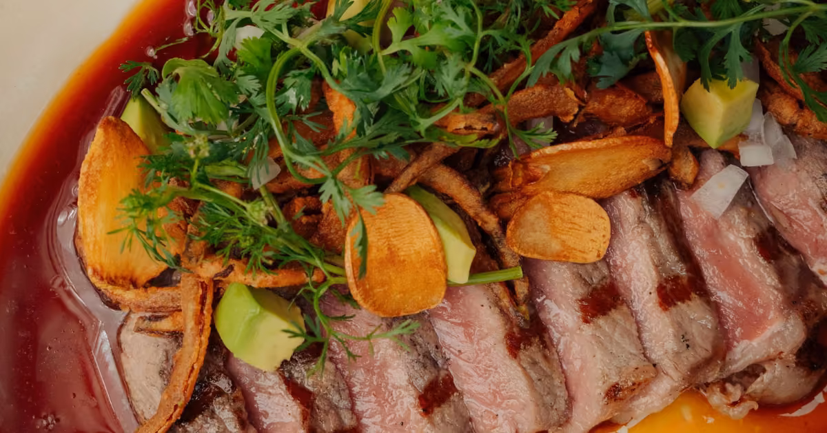 Grilled steak slices topped with fresh herbs, crispy garlic chips, and avocado, served with a rich red sauce.