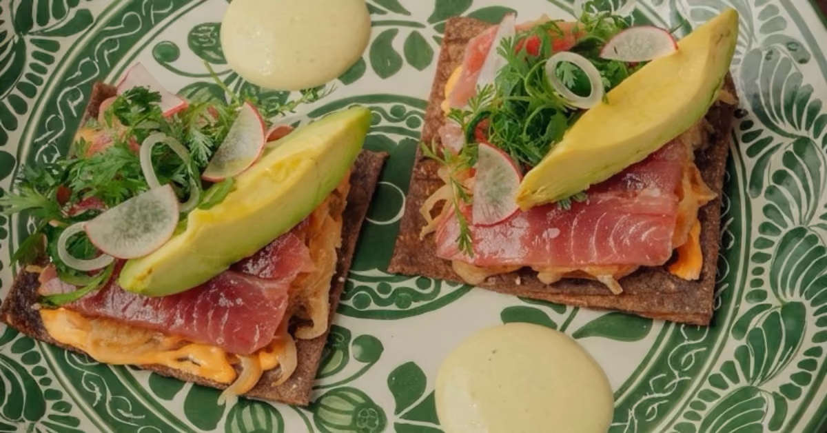 Flatbreads with raw tuna, avocado, radish, herbs, and creamy sauce on a green-white plate, extra sauce served beside.