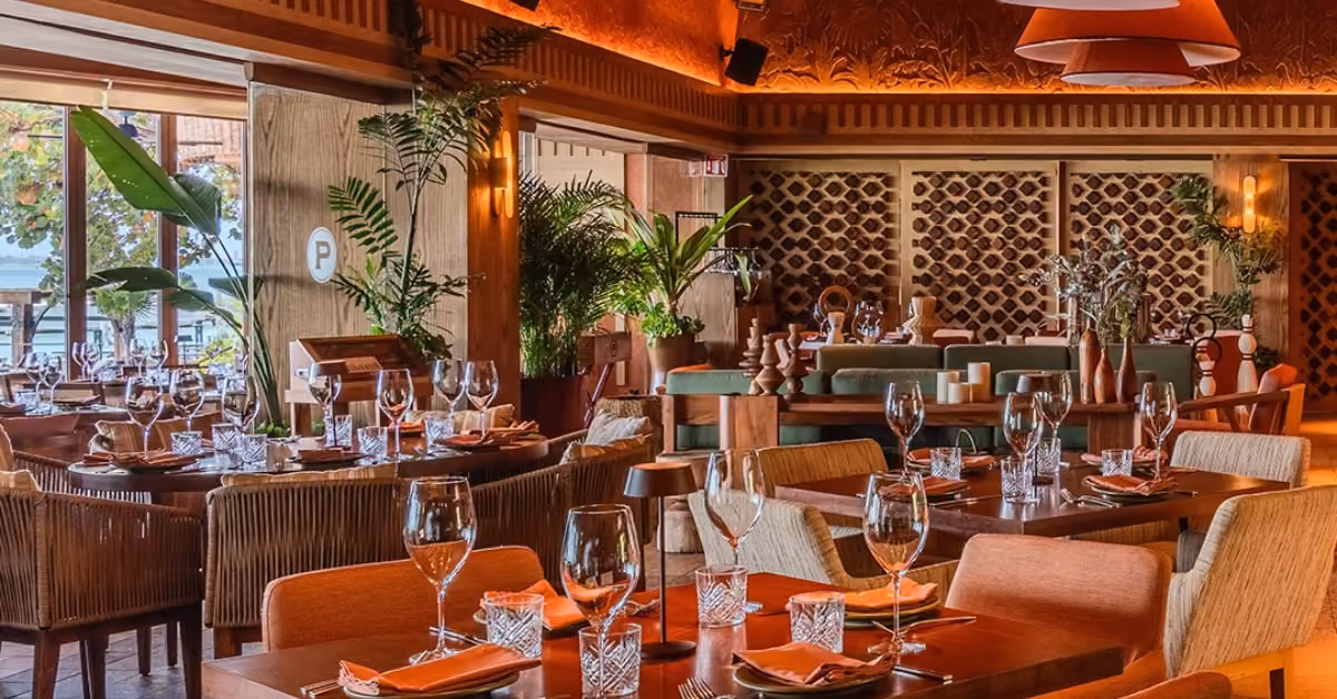 Porfirio’s restaurant with wooden tables, glassware, orange napkins, plants, warm lights, and large windows overlooking greenery.