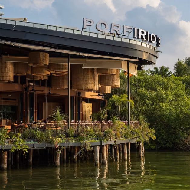 Porfirio’s sits on stilts above the water, surrounded by greenery and hanging lights, with tables under a partly cloudy sky.