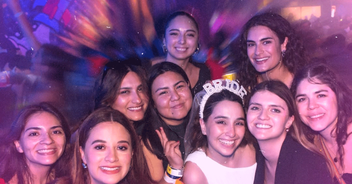 Eight women smile together at a lively party; one wears a BRIDE headband. Colorful lights add to the festive atmosphere.
