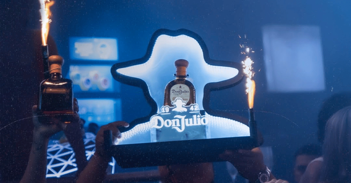 A Don Julio tequila bottle with sparklers is held up in a lively nightclub, blue lights and people in the background.