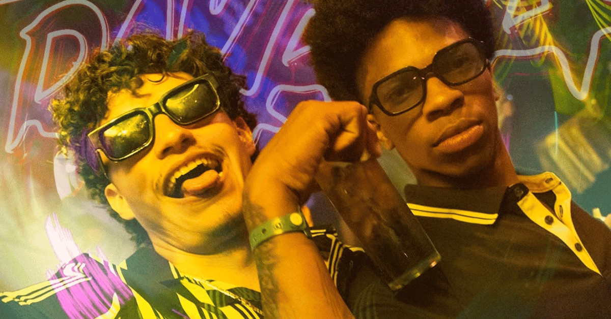 Two young men in sunglasses and striped shirts pose at a lively party; one holds a drink and sticks out his tongue amid neon lights.