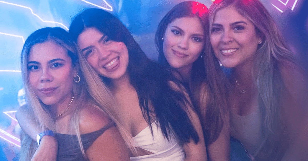 Four young women smile and pose together at Rakata Night Club, lit by blue and neon lights in trendy party outfits.