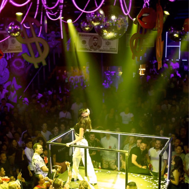 A woman stands in a spotlight on a platform in a lively nightclub with disco balls and dollar signs as the crowd watches below.