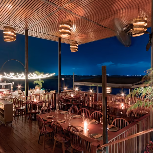 Cozy outdoor restaurant in Cancun at night, wooden tables, warm hanging lamps, and views of the dark sky over shimmering water.