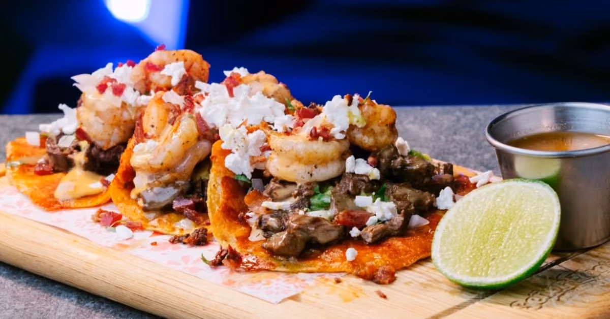 Three tacos with shrimp, steak, cheese, herbs, and bacon on a wooden board, served with lime and sauce at Señor Frog's.