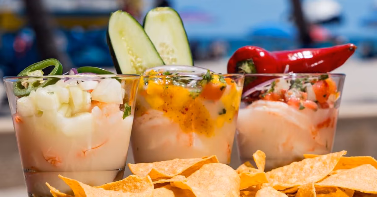 Glasses of shrimp ceviche with cucumber, mango, and chili sit behind tortilla chips, beach blurred in the background.