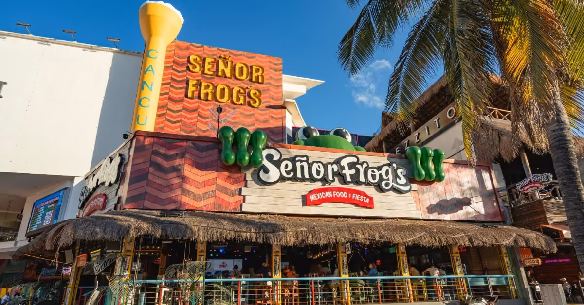 Señor Frog’s Cancun exterior with colorful signs, a yellow cup, frog eyes above entrance, palm tree, and people nearby.
