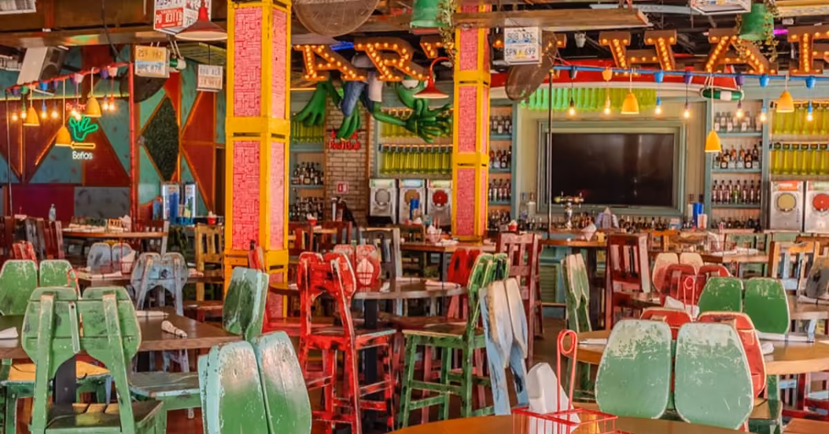 Bright Señor Frog's interior with mismatched chairs, colorful walls, neon signs, and a lively vibe. Bar and TV in back.