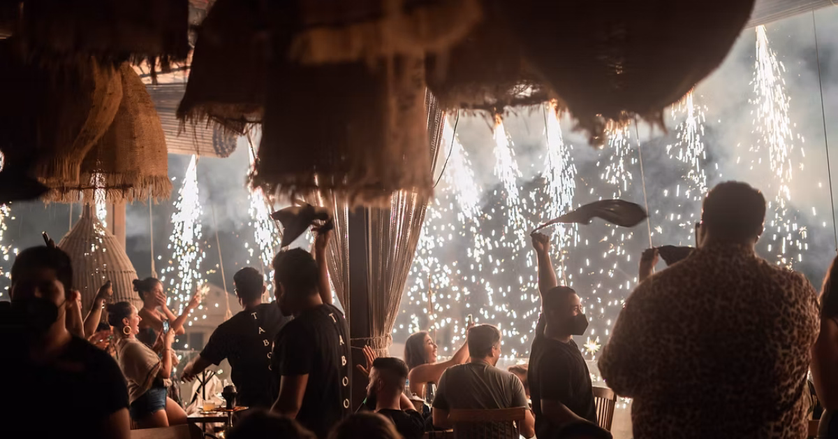 People sit and stand at a lively restaurant party, some raising hands as sparklers light up the festive, vibrant background.