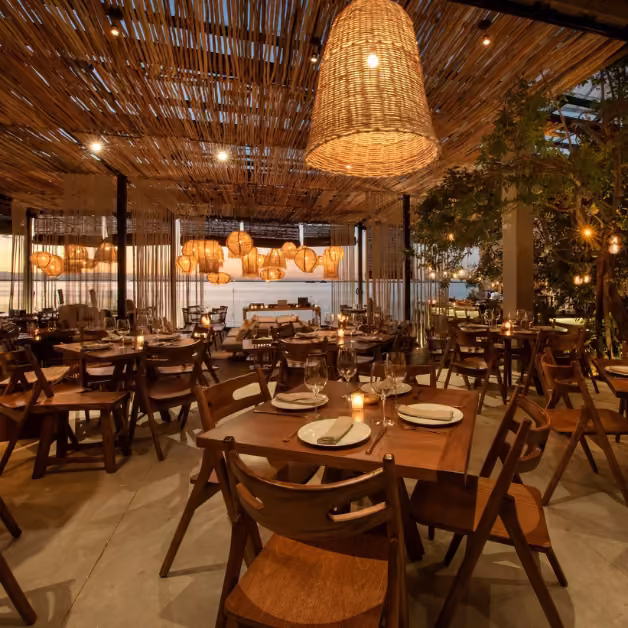 Cozy restaurant with wooden tables, chairs, wicker lights, and a woven ceiling; tables set for a warm, inviting meal.