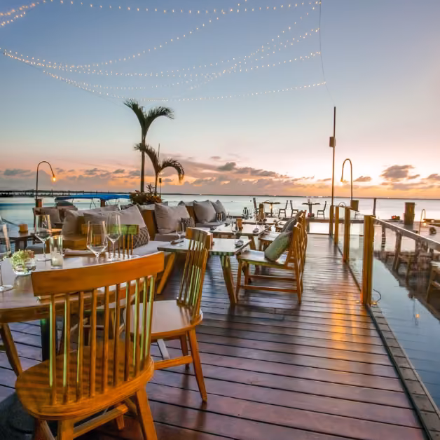 Outdoor restaurant deck with wooden tables, cushioned chairs, string lights, and palm trees overlooking the ocean at sunset.