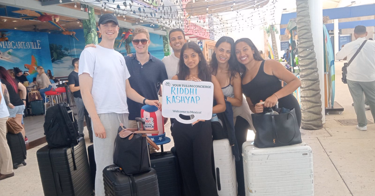 Six people smile with luggage at an airport terminal. One holds a “Riddhi Kashyap - Welcome to Mexico” sign; palm trees behind.