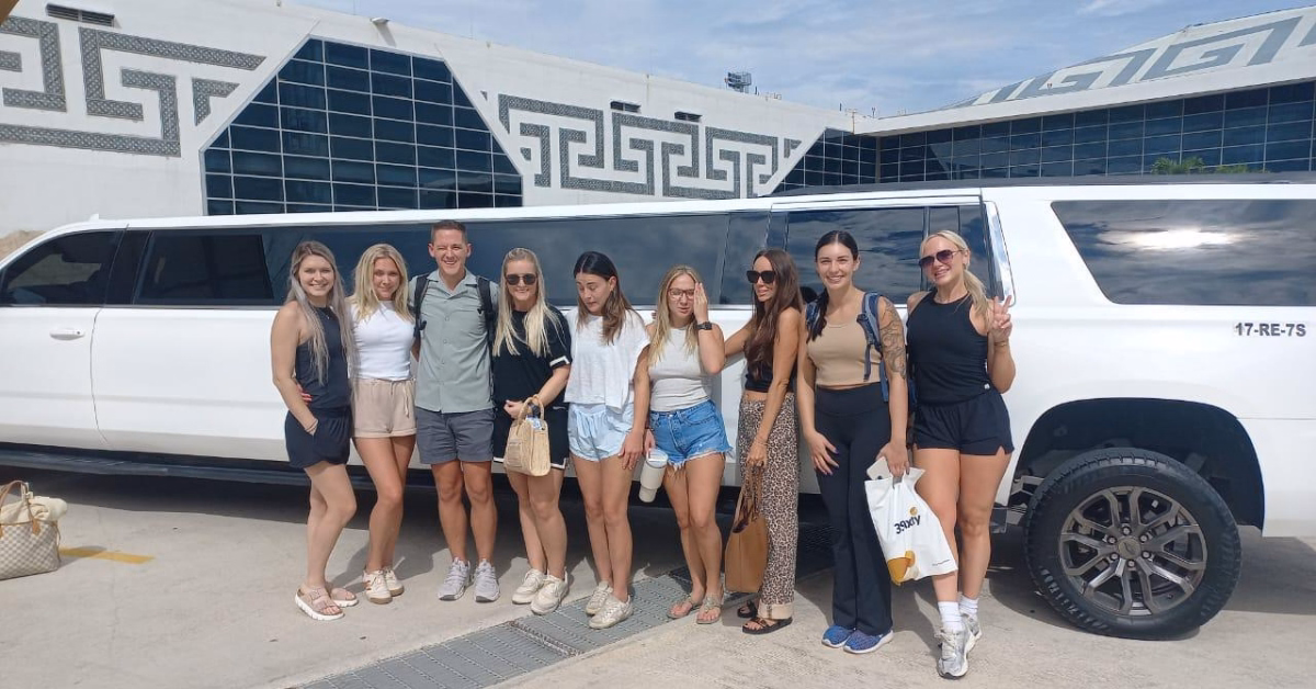 Eight people—seven women and one man—smile in front of a white limo parked outside a modern building with geometric designs.