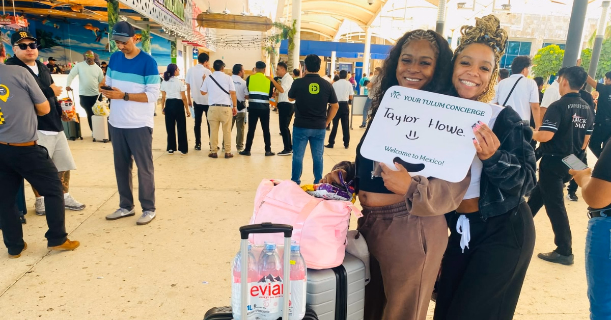 Two women at an airport smile and hold a welcome sign by their luggage, with others waiting in the background.