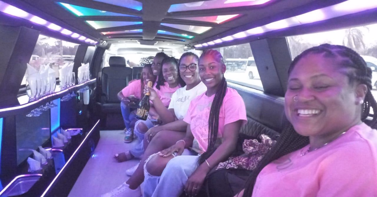 Five women in pink shirts smile and pose inside a brightly lit limo with colorful lights and drinks along the side.