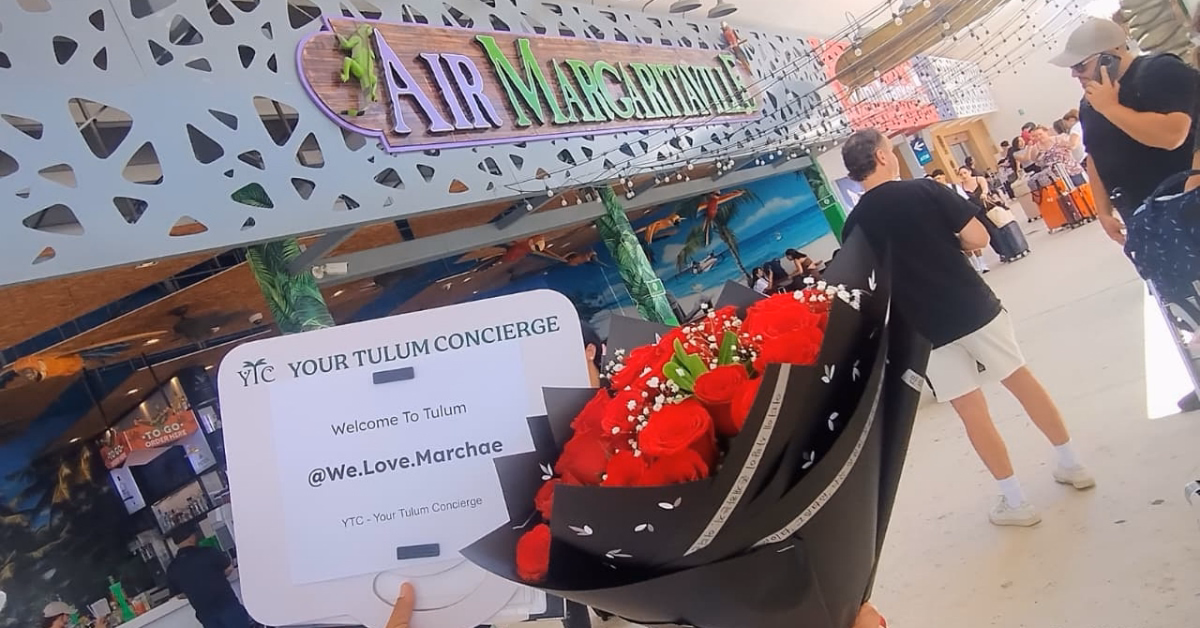 A person with red roses and a welcome sign waits near an Air Margaritaville sign at the airport, others in the background.