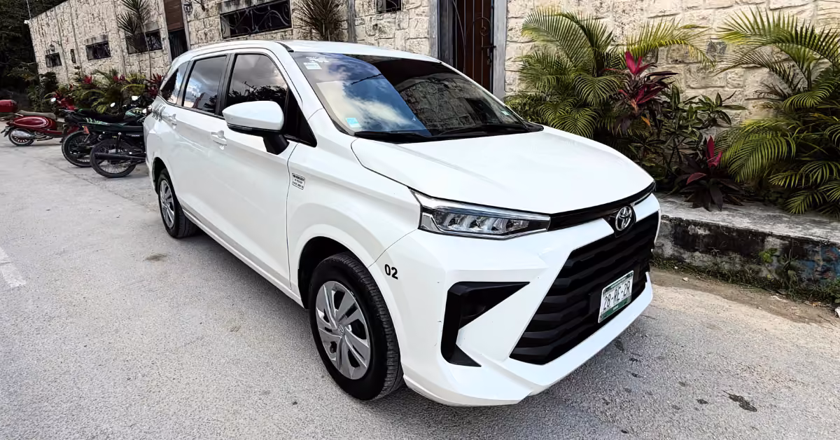 A white Toyota Avanza is parked on a street next to a stone wall with green and red plants, facing left with plate visible.