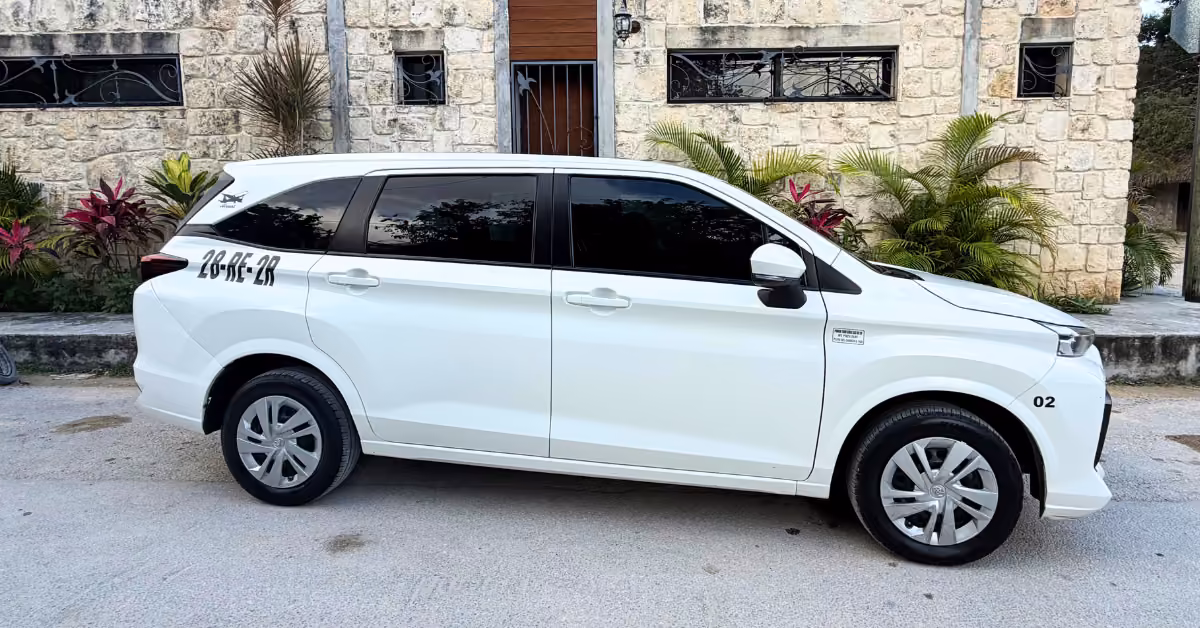A white minivan with tinted windows is parked by a stone building with plants. Numbers 28-RE-2R and 02 are on the vehicle.