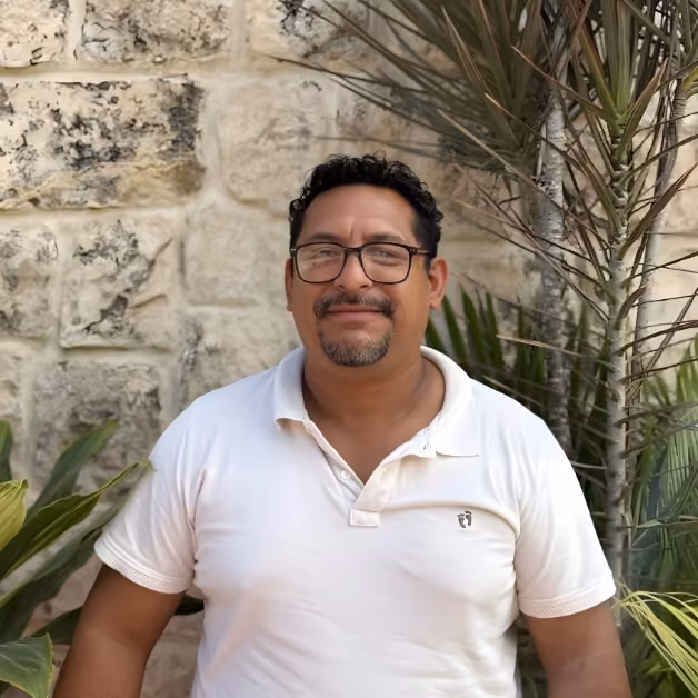 Smiling man with short curly hair, glasses, and a goatee stands by a stone wall and tropical plants in a white polo shirt.