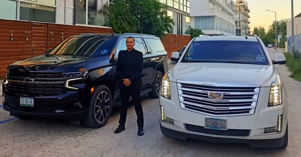 A driver in a black suit stands between a black Chevrolet and a white Cadillac SUV parked by modern buildings and a wooden fence.