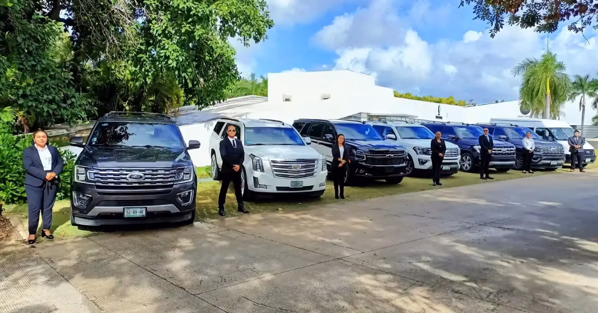 Seven people in formal wear stand by SUVs and luxury cars with their driver, near trees and a white building on a sunny day.