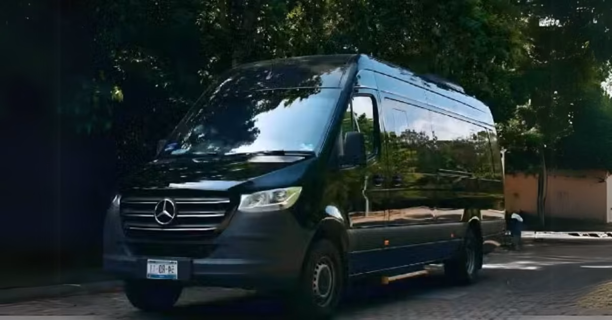A black Mercedes-Benz Sprinter van, used as an airport shuttle, is parked on a shaded street with trees and sunlight reflecting.