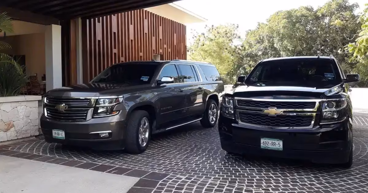 Two black Chevrolet SUVs with a driver are parked side by side on a driveway by a modern building with wood panels and greenery.