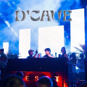 A DJ plays on stage, blue lights and LED screens glowing behind them. “DCAVE” shines above as the crowd watches below.