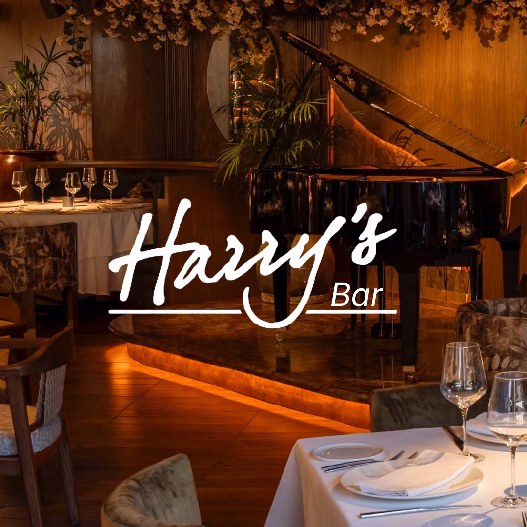 Cozy, upscale restaurant with a grand piano, set tables topped with wine glasses, and Harrys Bar in elegant white script.